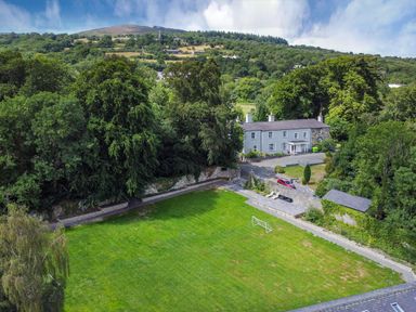 A large green lawn with a small soccer goal near a stone wall and a gray house surrounded by trees at Bryn Derwen in Bethesda near Caernarfon