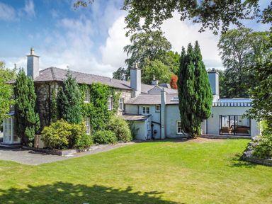 An exterior view of a large stone and white two-story house with multiple chimneys surrounded by tall trees and a lawn at Bryn Derwen Bethesda near Caernarfon