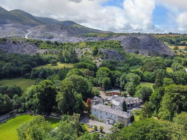 An aerial view of a large house surrounded by trees and fields with rocky hills in the background at Bryn Derwen in Bethesda near Caernarfon