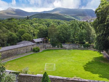 A green open field with two small soccer goals surrounded by stone walls and trees with mountains in the background at Bryn Derwen in Bethesda near Caernarfon