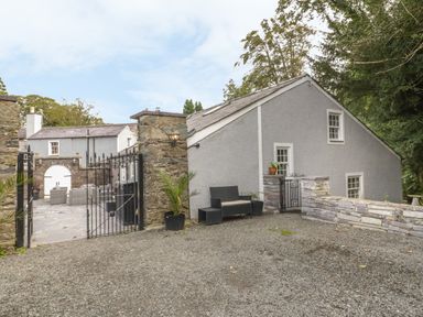 An outdoor stone patio with seating and potted plants next to a grey building with white framed windows at Bryn Derwen Coach House in Bethesda