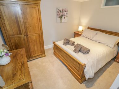A bedroom with a wooden bed frame and wardrobe a bedside table with a lamp and a chest of drawers with flowers at Bryn Derwen Coach House in Bethesda