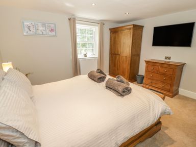 A bedroom with a bed white bedding rolled towels wooden wardrobe chest of drawers and a wall mounted tv at Bryn Derwen Coach House in Bethesda