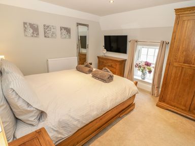 A bedroom with a wooden bed with pillows and towels a wooden wardrobe a chest of drawers a wall-mounted tv a mirror and a window with flowers at Bryn Derwen Coach House in Bethesda