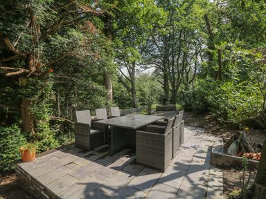 An outdoor patio area with a rectangular table and six wicker chairs surrounded by trees and greenery at Bryn Derwen Coach House in Bethesda