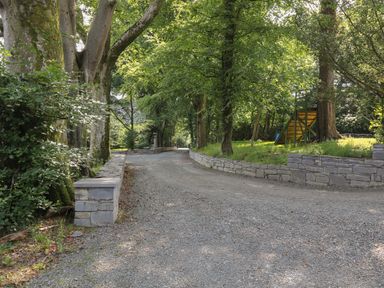 A gravel pathway lined with stone walls and large trees in a wooded area at Bryn Derwen Coach House in Bethesda
