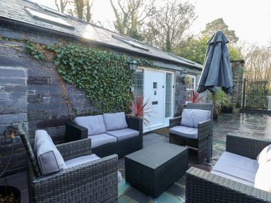 An outdoor patio with rattan furniture and cushions in front of a stone wall with ivy at Bryn Derwen Coach House in Bethesda