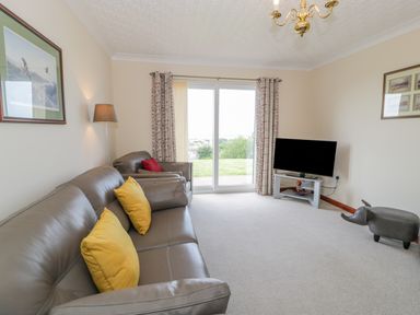 A living room with leather sofas and yellow cushions a television on a stand and glass sliding doors at Bryn Coch Bach in Dyffryn Ardudwy