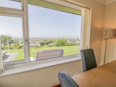 A dining table with chairs next to a large window showing a garden and distant houses at Bryn Coch Bach in Dyffryn Ardudwy
