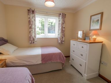 A bedroom with two single beds a window with floral curtains and a white dresser with a lamp and framed picture at Bryn Coch Bach in Dyffryn Ardudwy