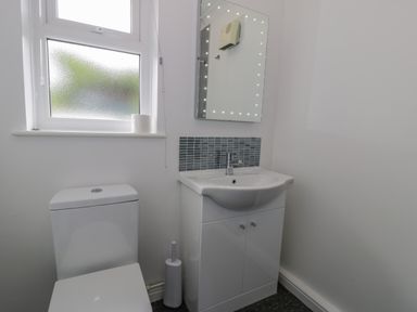 A bathroom with a toilet, a sink cabinet, a mirror with lights, a window, and a toilet brush at Bryn Coch Bach in Dyffryn Ardudwy