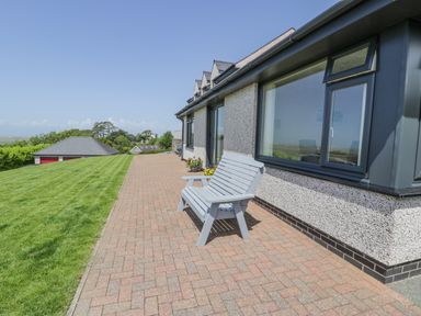 A bench on a brick patio next to windows of a house with a lawn and garage in the background at Bryn Coch Bach in Dyffryn Ardudwy