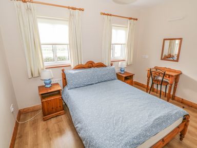 A bedroom with a wooden bed and blue bedspread two bedside tables with lamps two windows with curtains and a wooden dressing table with a chair at 8 Lios Na Sioga Binghamstown near Belmullet County Mayo