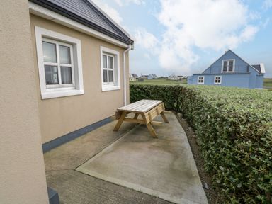 An outdoor patio area with a wooden picnic table beside a beige house and trimmed hedges at 8 Lios Na Sioga in Binghamstown near Belmullet County Mayo