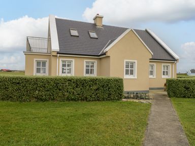 A beige house with white windows and a dark roof surrounded by green grass and hedges at 8 Lios Na Sioga in Binghamstown near Belmullet County Mayo