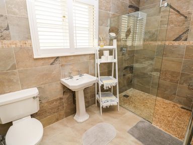 A bathroom with a toilet a pedestal sink a white shelving unit with towels and toilet paper a glass shower enclosure and tiled walls and floors at Llety'r Llwynog in Llanfallteg near Narberth