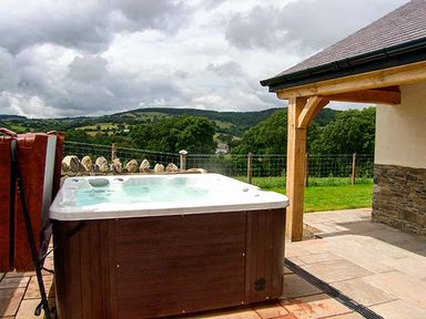 An outdoor hot tub on a stone patio next to a wooden porch with hills and trees in the background at Tyn Y Celyn Uchaf in Clocaenog near Ruthin