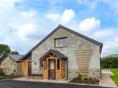 A detached house with a wooden porch and trellis on the wall surrounded by garden beds and a paved driveway at Tyn Y Celyn Uchaf in Clocaenog near Ruthin