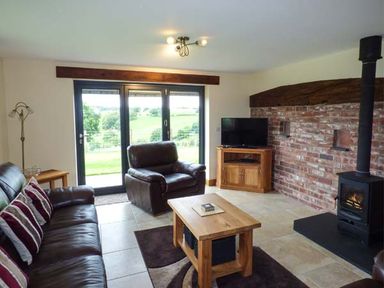 A living room with leather sofas a wooden coffee table a wood stove and a television with glass doors showing outside at Tyn Y Celyn Uchaf in Clocaenog near Ruthin