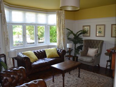 A sitting room with leather sofas, an armchair, a wooden coffee table, lamps, framed pictures, and a large window at Lily House in Minehead