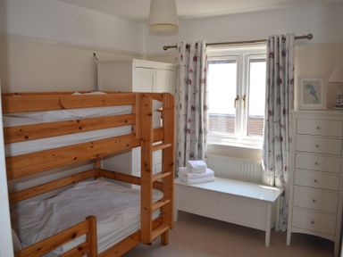 A bedroom with wooden bunk beds a white chest of drawers a white storage bench with towels and a window with patterned curtains at Lily House Minehead