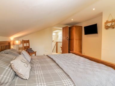 A bedroom with a bed, pillows, a chair with folded towels, a wooden wardrobe, a wall-mounted TV, and a lamp at Higher Whiddon Farm Whiddon Well Ashburton near Newton Abbot