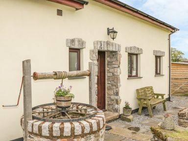 An outdoor area with a stone well, wooden bench, potted plants, stone walls, and a door at Higher Whiddon Farm Whiddon Well Ashburton near Newton Abbot