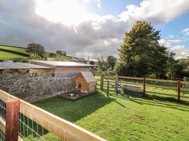 A fenced grassy yard with a wooden dog house a stone wall and trees at Higher Whiddon Farm Whiddon Well Ashburton near Newton Abbot