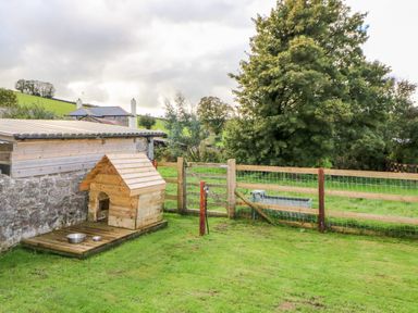 A wooden dog house with two metal bowls on a wooden platform next to a stone and wood shed in a fenced grassy yard at Higher Whiddon Farm Whiddon Well Ashburton near Newton Abbot