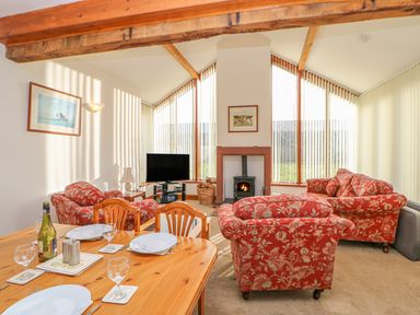 A living room with red floral armchairs a wooden dining table and chairs a TV a fireplace and large windows at Swift Cottage in Milton near Brampton