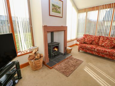 A living room with a wood burning stove a basket of firewood a red patterned sofa and windows with vertical blinds at Swift Cottage in Milton near Brampton