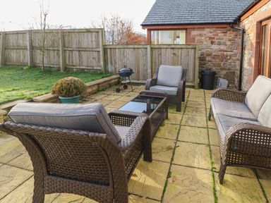 An outdoor patio with three wicker chairs a wicker sofa a glass coffee table a small barbecue grill potted plant wooden fence and stone wall at Swift Cottage Milton near Brampton