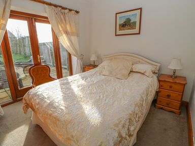 A bedroom with a bed between two wooden bedside tables and lamps and a chair near glass doors at Swift Cottage in Milton near Brampton