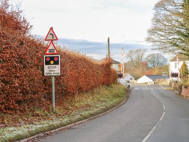 A road with traffic signs indicating a left turn and a level crossing with stop lights at Swift Cottage in Milton near Brampton