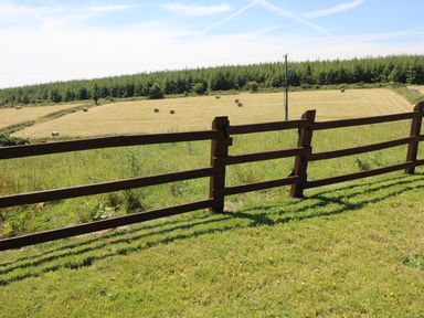 A wooden fence with green grass in front of fields with hay bales and trees in the background at Clash-Brack in Dungarvan County Waterford