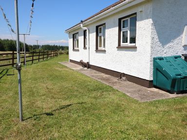 A side view of a white house with three windows a green bin and a grassy yard with a fence in the background at Clash-Brack in Dungarvan County Waterford