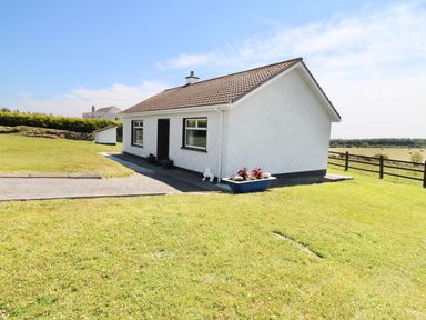A small white house with a tiled roof on a grassy lawn with a planter and a wooden fence at Clash-Brack in Dungarvan County Waterford