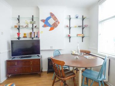 A dining area with a round wooden table and mixed wooden chairs next to a TV on a wooden cabinet and wall shelves at Tidewood in Polzeath