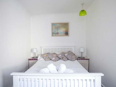 A bedroom with a white bed framed by two bedside tables with lamps and a painting on the wall at Tidewood in Polzeath