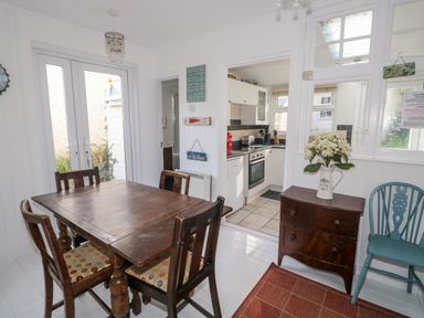 A dining area with a wooden table and four chairs next to a kitchen with white cabinets and appliances at Driftwood Polzeath near Wadebridge