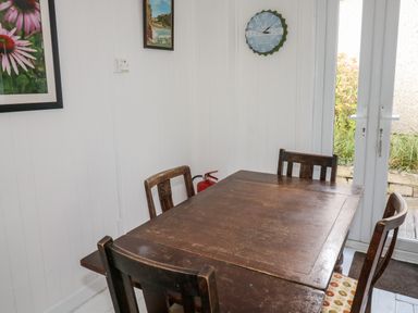 A dining area with a wooden table and four chairs beside white walls with pictures and a clock at Driftwood Polzeath near Wadebridge