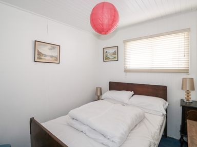A bedroom with white walls a double bed white bedding two bedside tables with lamps paintings on the wall and a red ceiling light at Driftwood in Polzeath near Wadebridge