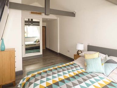 A bedroom with a bed covered in a geometric pattern bedspread and pillows a wooden chest of drawers with a blue vase a bedside table with a lamp and a mirror on the wall at White Cottage Annexe in Tenby