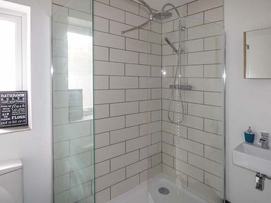 A bathroom with a corner shower with glass walls a tiled wall a small white sink and a window with a signs at White Cottage Annexe in Tenby