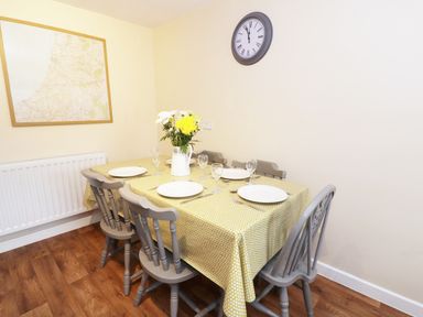 A dining table with six chairs set with plates glasses and cutlery a vase with flowers on the table a wall clock and a framed map on the walls a radiator under the map at Penglanowen Fawr in Llangwyryfon near Aberystwyth