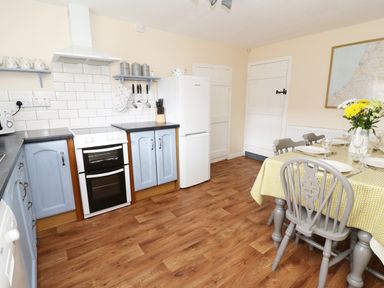 A kitchen with blue cabinets a white stove and oven a white refrigerator a dining table with yellow tablecloth and gray chairs with flowers on the table at Penglanowen Fawr in Llangwyryfon near Aberystwyth