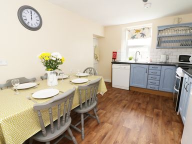 A kitchen with blue cabinets a dining table with chairs and flowers a wall clock and appliances at Penglanowen Fawr in Llangwyryfon near Aberystwyth