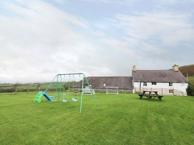 A playground with swings and a slide in a grassy yard with picnic tables near a white cottage at Penglanowen Fawr in Llangwyryfon near Aberystwyth