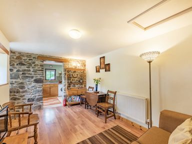 A room with wooden chairs a small wooden table a wooden shelf a floor lamp and a stone wall leading to a kitchen at Cilwen Fach in Cynwyl Elfed