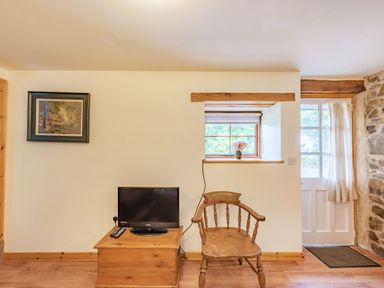 A wooden chair next to a small TV on a wooden table under a window with a flower vase and a door with curtains at Cilwen Fach in Cynwyl Elfed